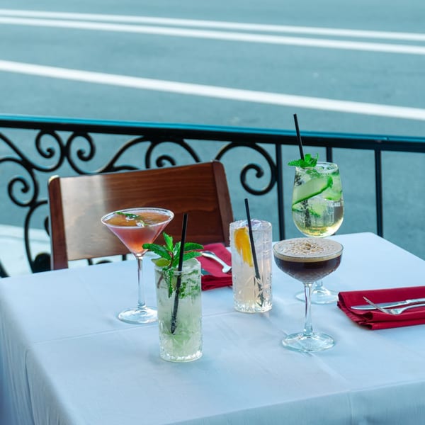 Five colorful cocktails on a white-clothed table on a restaurant patio overlooking a street.