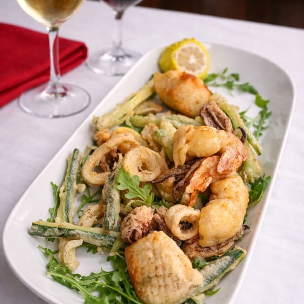 Fried seafood platter with calamari, shrimp, and zucchini, garnished with arugula and lemon, alongside wine glasses.