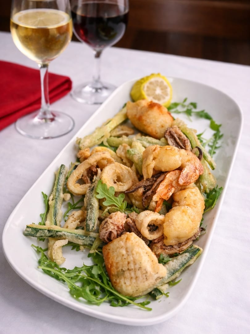 Fried seafood platter with calamari, shrimp, and zucchini, garnished with arugula and lemon, alongside wine glasses.