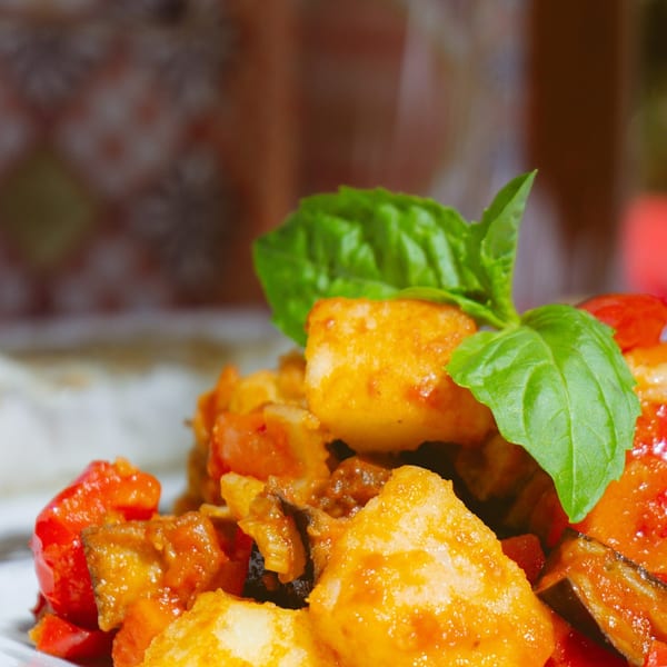 Close-up of golden fried potatoes, red peppers, and eggplant, garnished with fresh basil on a white plate.