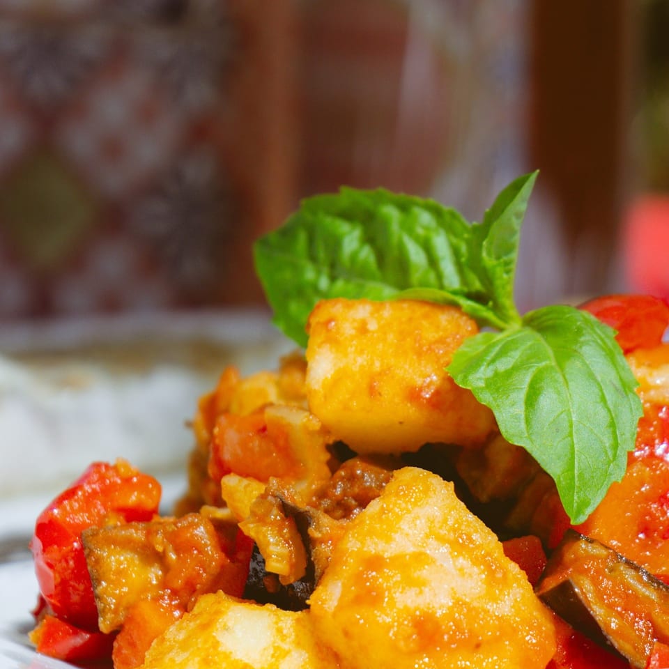 Close-up of golden fried potatoes, red peppers, and eggplant, garnished with fresh basil on a white plate.
