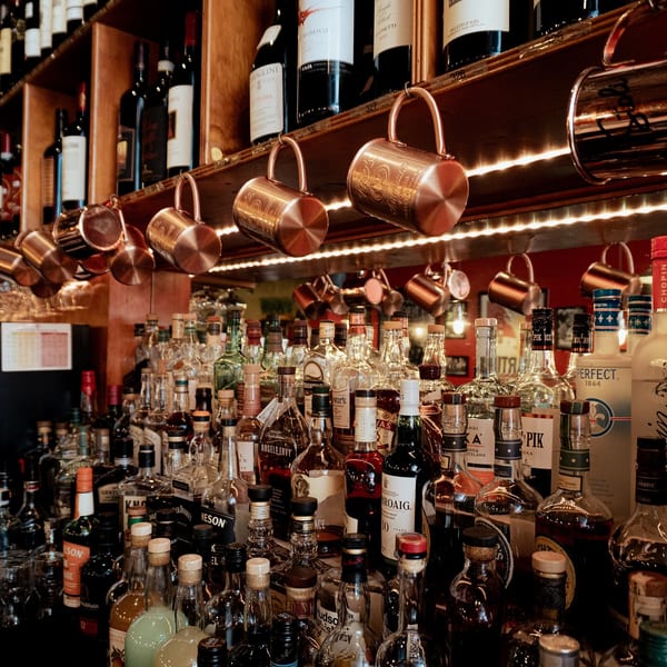 A well-stocked bar with rows of liquor bottles, wine bottles on shelves, and copper mugs hanging above.