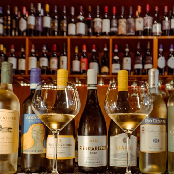 Two wine glasses filled with white wine sit on a bar, surrounded by various wine bottles.
