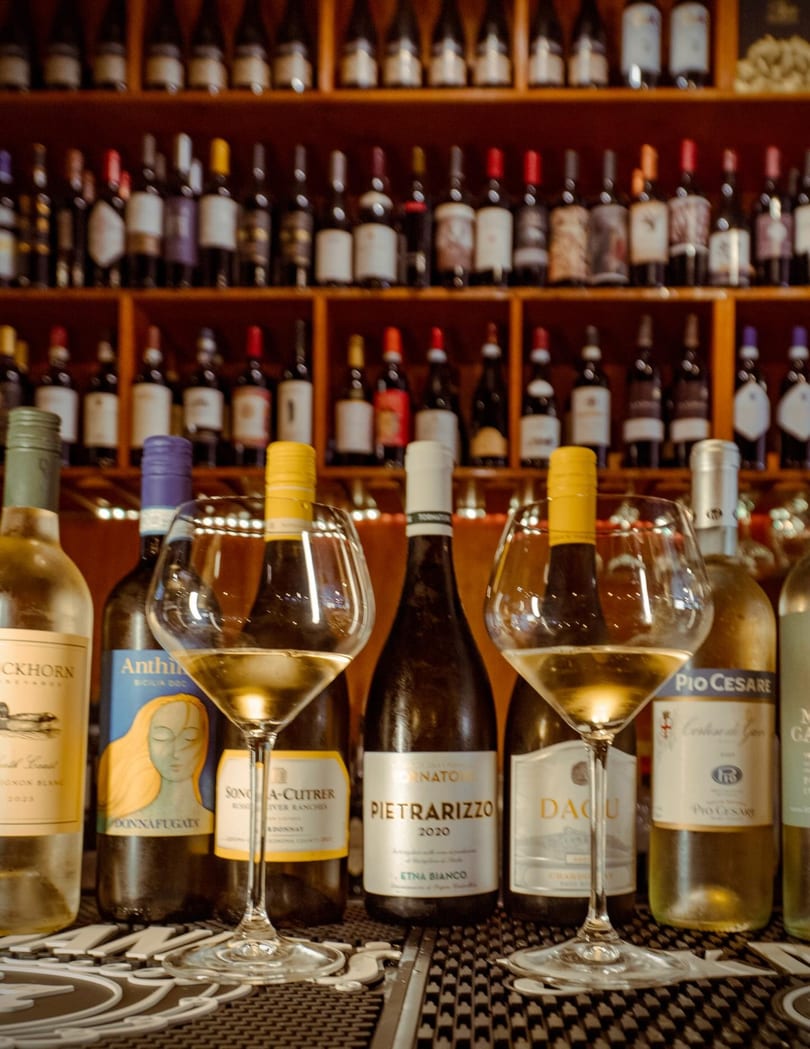 Two wine glasses filled with white wine sit on a bar, surrounded by various wine bottles.
