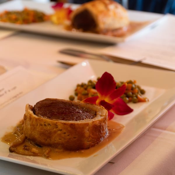 Beef Wellington with gravy, peas, carrots, and a red orchid on a white plate.