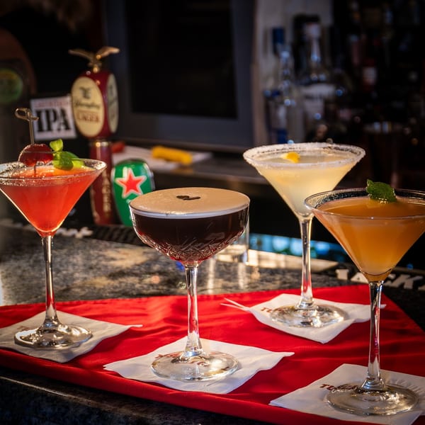 Four colorful cocktails, including an espresso martini, on a red tray at a bar.