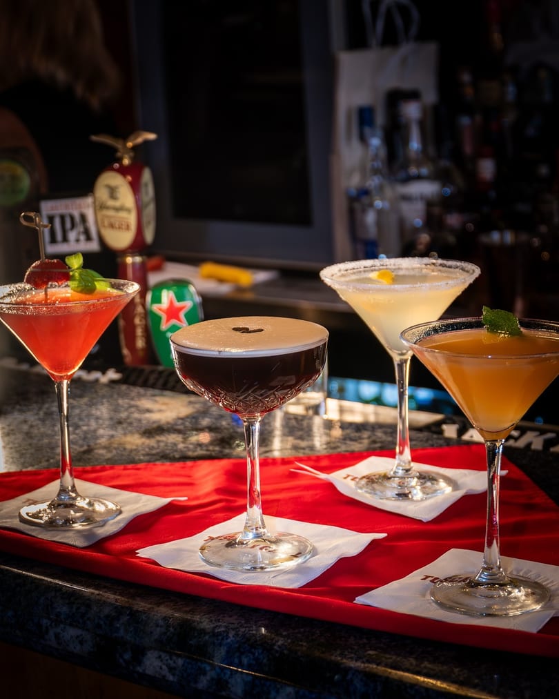 Four colorful cocktails, including an espresso martini, on a red tray at a bar.