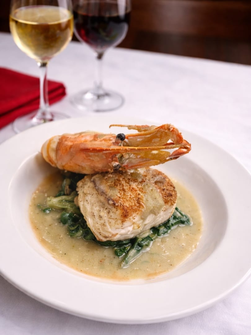 Pan-seared fish with a large prawn, spinach, and sauce, served with wine glasses.