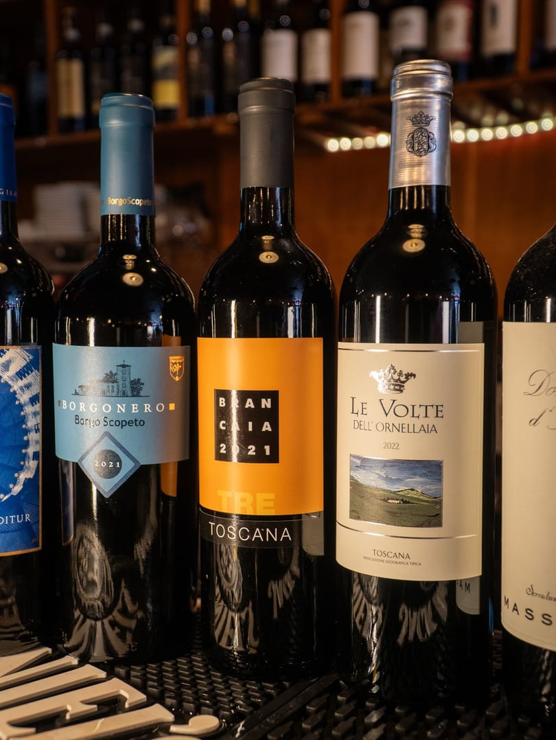 Five dark wine bottles with colorful labels, including "Borgonero" and "Le Volte," stand on a bar.